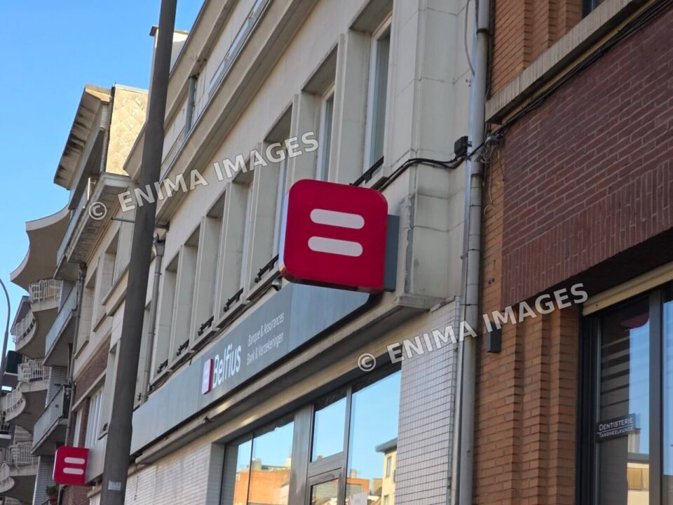 Façade d’agence Belfius vue en perspective avec deux logos rouges, des fenêtres et une façade voisine en briques.