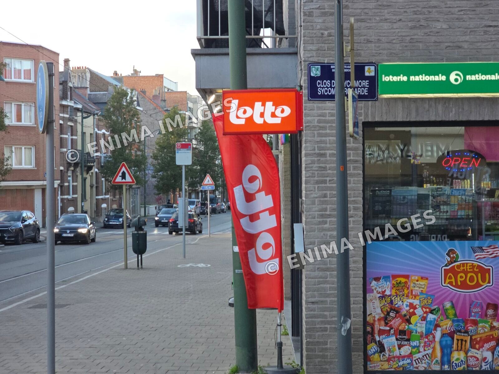 Vitrine d’un commerce portant une enseigne Lotto orange, un drapeau rouge et un bandeau vert de la Loterie Nationale, au bord d’une rue parcourue par des rails de tram.