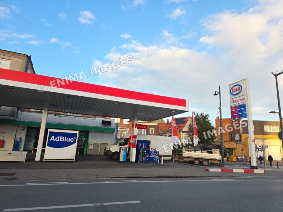 Station-service Esso Express sous un auvent rouge et blanc, avec des pompes Synergy, un équipement AdBlue et un totem de prix. Une camionnette et une remorque sont visibles.