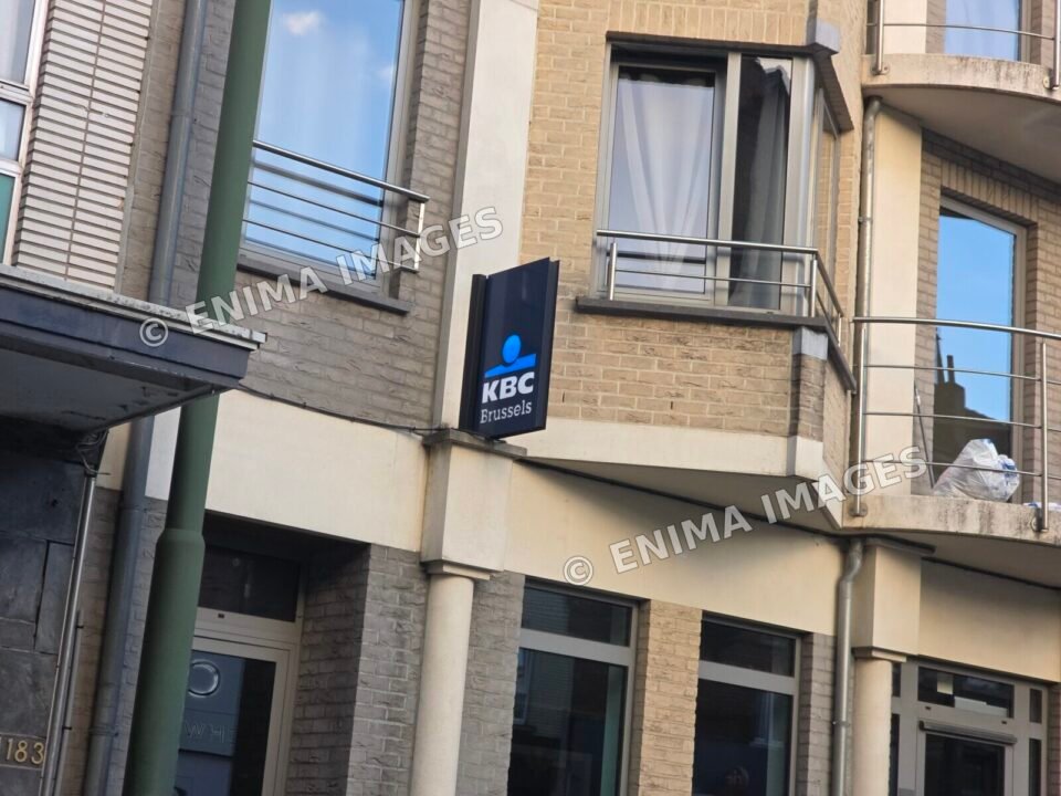 Vue élargie d’une façade en briques claires portant une enseigne KBC Brussels, avec les vitrages du rez-de-chaussée, des fenêtres et des balcons aux étages.