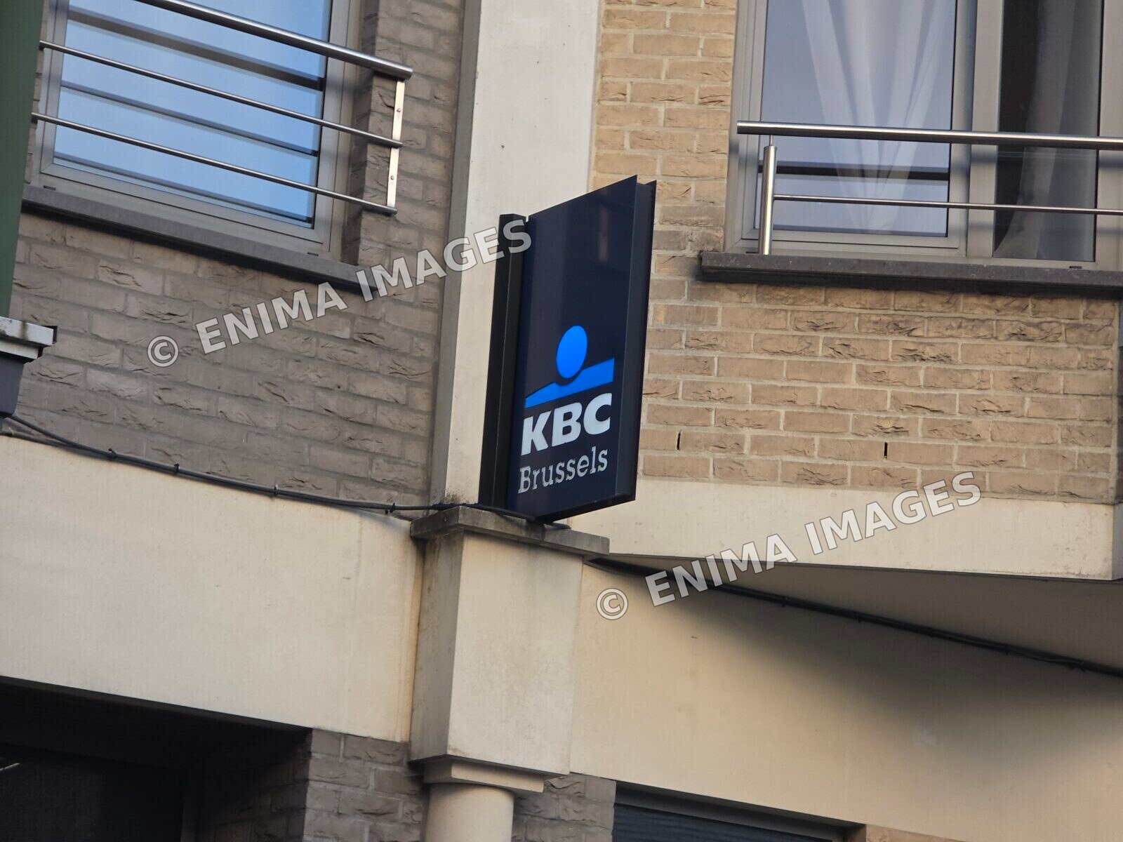 Enseigne noire KBC Brussels au logo bleu, fixée perpendiculairement à une façade en briques claires sous des fenêtres avec garde-corps métalliques.